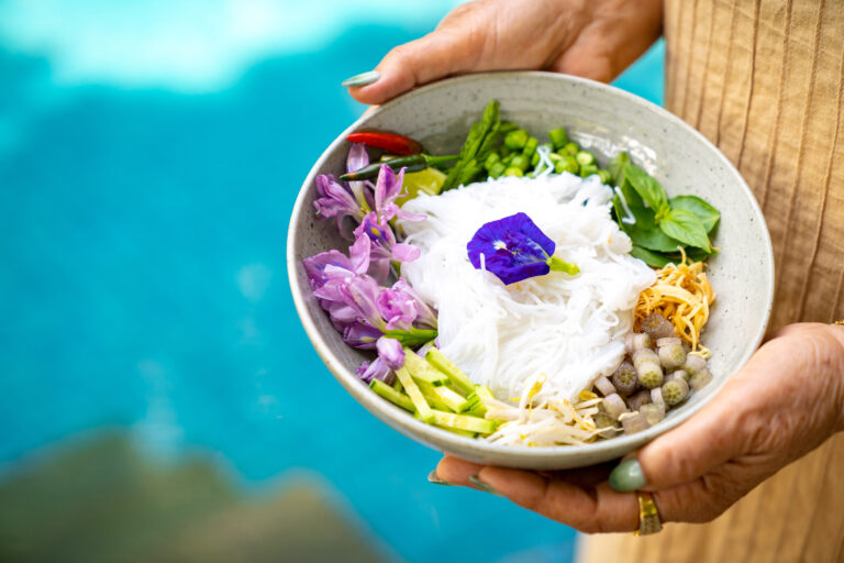 Bowl of noodles with fresh herbs, bean sprouts, and edible flower garnish